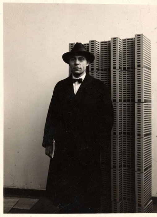 En man i hatt och rock står framför en modell av höghus, i en vintage svartvit bild.