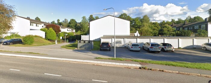 Flerfamiljshus i vitt med parkerade bilar i förgrunden och omgivande grönska, typisk förortenmiljö.