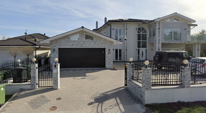 Stor villa med stenfasad, stor garageport och inhägnad gårdsplan. Bilar parkerade framför huset.