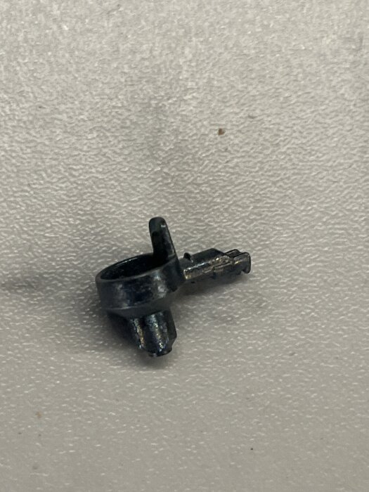 En svart metallplugg med en böjd topp och ojämn yta, ligger på en ljusgrå yta.