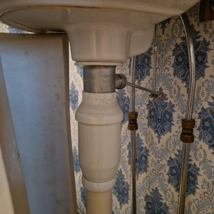 Under diskbänk med vattenlås och rör på väggen, blått blommigt tapetmönster i bakgrunden.