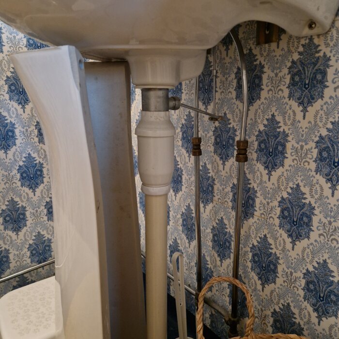 Handfat med avloppsrör och vattenlås mot blåmönstrad tapet i ett badrum.