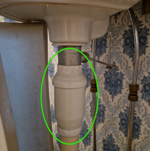 Vattenlås under ett handfat, omgivet av rör och mönstrad tapet i bakgrunden.