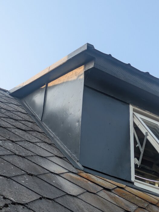 Ovanvåningens takkupa i plåt med öppet fönster, visar takkonstruktion med skifferplattor under blå himmel.
