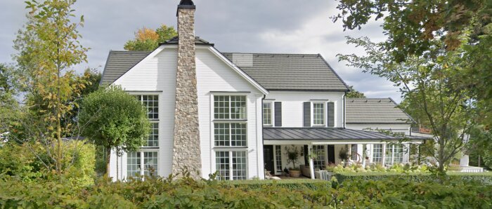 Vit villa med stenväggad skorsten, stora fönsterpartier och svart tak, omgiven av träd och grönska, på Lidingö.