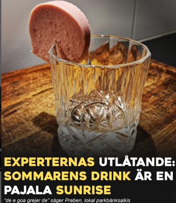 Ett tomt glas med en skiva falukorv på kanten, på en träyta, med text som säger att sommarens drink är Pajala Sunrise.