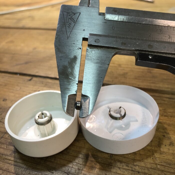 Skjutmått mäter djupet på hålet i en rund vit dimmerknapp, där två knappar syns liggande på ett träbord.