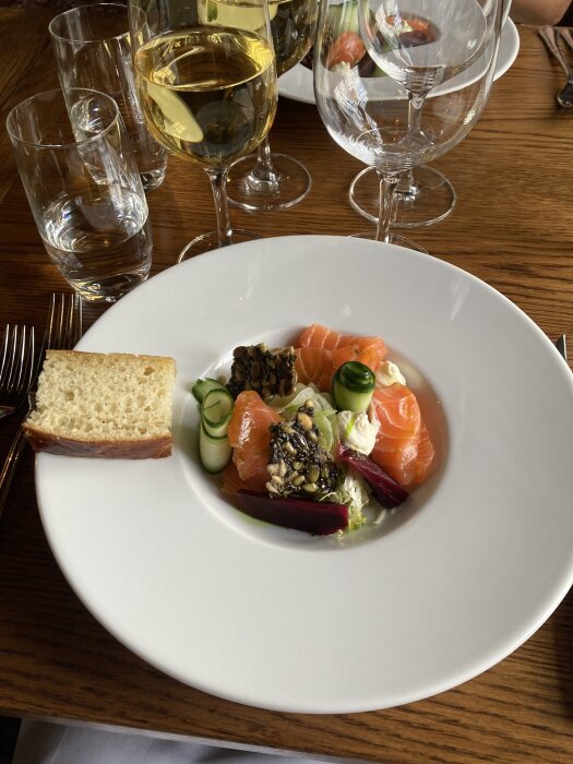 Elegant tallrik med laxrätt, bröd och sallad. Två glas vin och tre vattenglas på ett träbord vid Mauritzbergs slott under golfweekend.