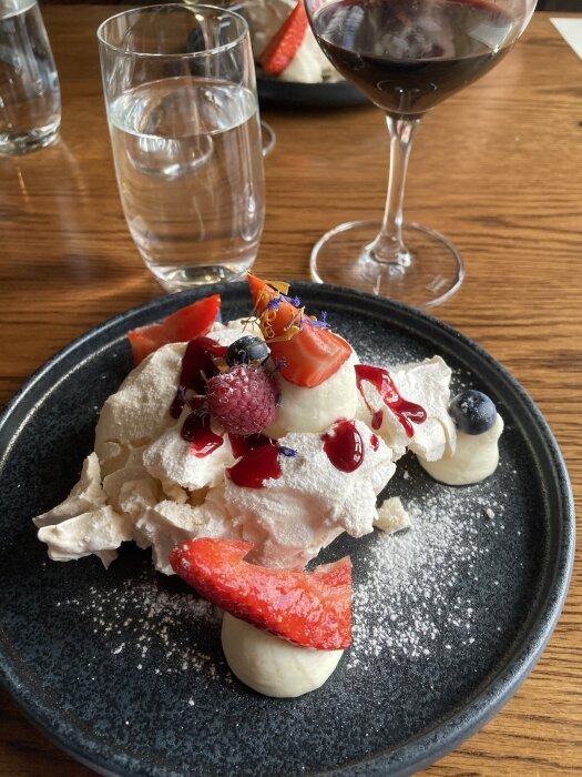 En elegant dessert med maräng, färska bär och grädde på ett svart fat, serveras med ett glas rött vin och vatten.