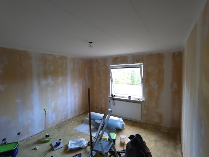 Rum under renovering med spacklade väggar, öppet fönster och byggverktyg på golvet.