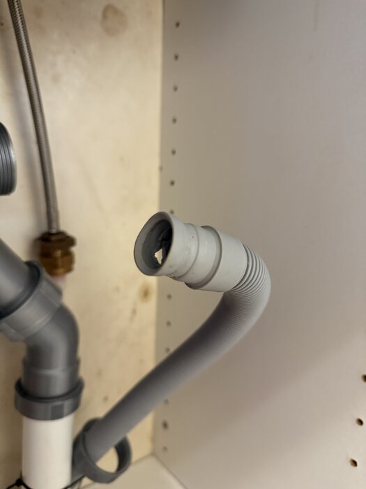 Grå avloppsslang i plast med skadat ändparti. Kopplingar och rör syns i bakgrunden, belägen under diskbänk.