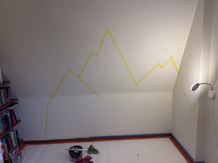 Vägg i barnrum med gul tejp i form av berg, lampa till höger, och böcker på en hylla till vänster. Golvet är täckt vid målning.