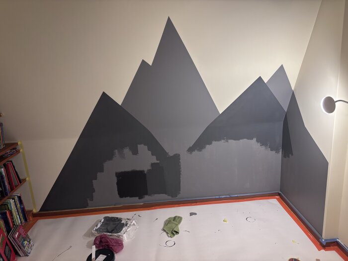 Väggmålning av berg i grått och svart under arbete i ett rum, med målartillbehör och skyddspapper på golvet.