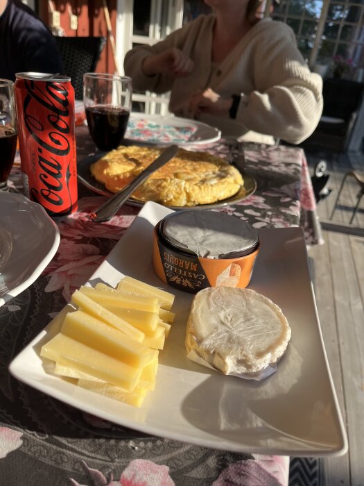 Tallrik med ostar och kex, en tortilla i bakgrunden, samt en burk Coca-Cola på ett dukat bord i solen.