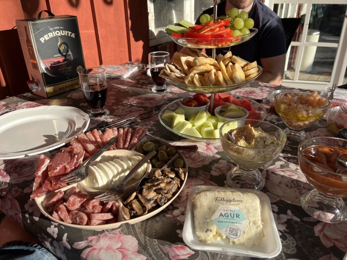 Tapasbord med ostar, kallskuret, frukt och bröd på blommig duk. Vinbox och glas på bordet.