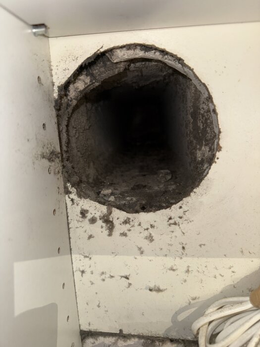 En rund öppning i en vägg med smutsig kant, troligen en ventilationskanal, omgivet av vit yta och delvis synliga kablar.