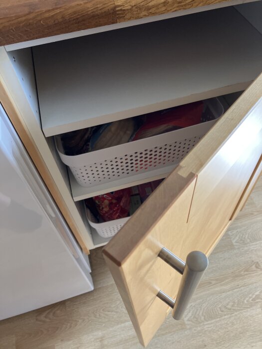 Öppet köksskåp med trähylla och en vit plastkorg fylld med förpackningar bakom en trälucka med metallhandtag.