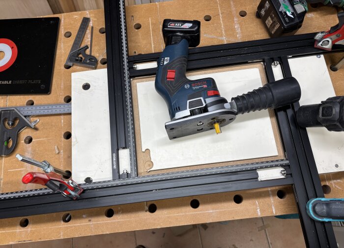 En Bosch trimmer med 12V batteri placerad på ett arbetsbord med mätverktyg och klämmor, omgiven av trä och tillbehör.