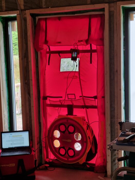 Provtryckningsutrustning installerad i dörröppning med röd tätning och fläkt. Laptop synlig till vänster.