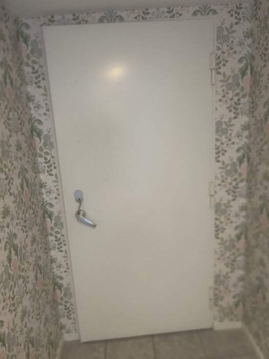 Vit ståldörr med låshandtag i en tapetserad korridor, där överkantens gångjärn orsakar friktion vid öppning.
