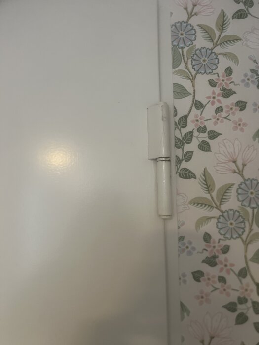 Gångjärn på en stor vit ståldörr mot blommigt tapetserad vägg. Justering behövs då dörren går emot vid överkanten.