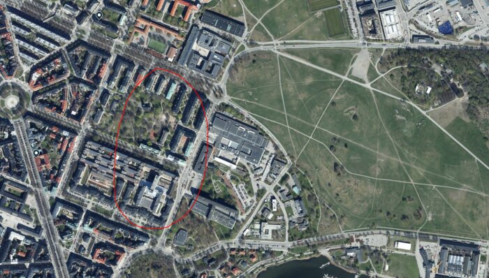 Flygfoto av ett stadsområde med byggnader inringade i rött, bredvid en stor park med gångvägar, troligen nära t-banestation.