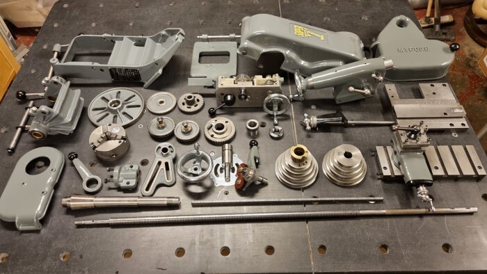 Delar av en svarv liggandes på ett bord efter demontering, inklusive kugghjul, axlar, och motorhus, alla målade i grått, redo för montering och användning.