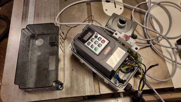 Verkstadens steg för installation av frekvensomriktare, med kablage och kontrollbox invid en svart plasthölje på träbord.