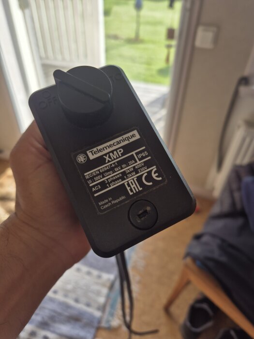 Tryckventil av märket Telemecanique XMP med streckade specifikationer, hålls i handen inomhus med trädgårdens ljus i bakgrunden.