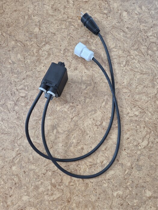 En tryckventil med två kablar, en med stickpropp och en med vit kontakt, ligger på ett mönstrat underlag.