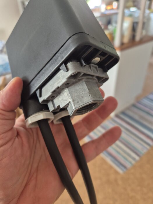 Hand håller en tryckventil med två anslutna kablar, inomhusmiljö i bakgrunden.