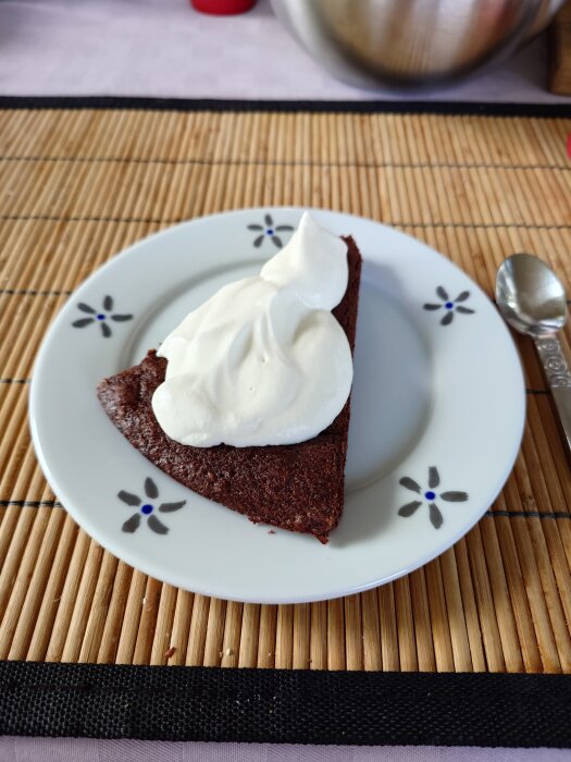En bit kladdkaka med vispad grädde på ett blomdekorerat fat, placerad på en bordstablett av bambu.