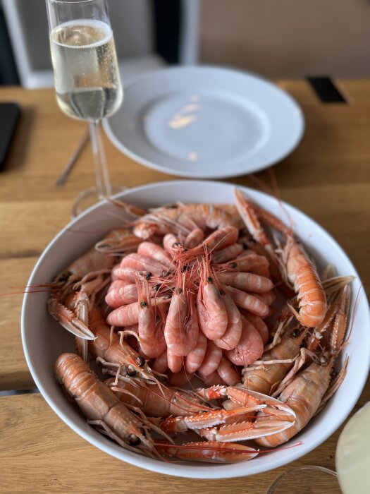 En skål med räkor och kräftor serverad på ett träbord, med ett glas mousserande dryck och en tom tallrik i bakgrunden.