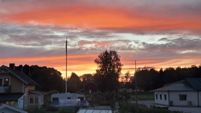 Solnedgång över ett landskap med hus, träd och flaggstång, himlen färgad i orange och rosa nyanser.