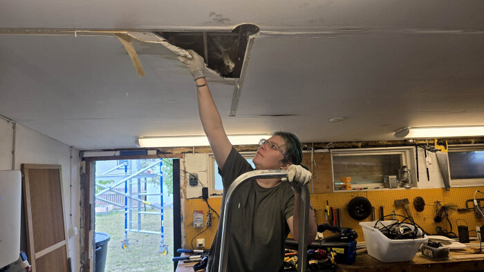 Person på stege inspekterar hål i garagetakets innertak, omgiven av verktyg och byggmaterial.