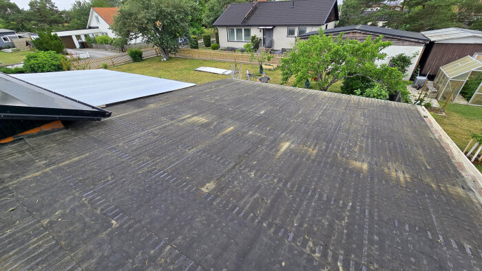 Platt tak med gammal takpapp och utsikt över närliggande villor och trädgårdar, pågående renovering av garagetak.