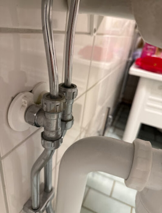 Vattenkopplingar och rör under ett handfat, med två metallanslutningar monterade på en kaklad vägg i ett badrum.