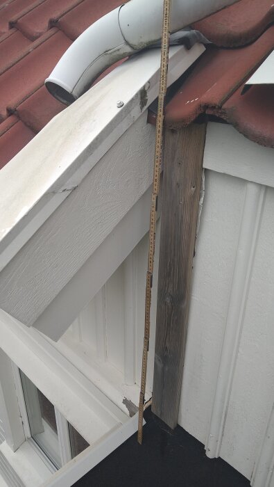 Mätstock vid takkupa med avloppsrör, plankor och röda takpannor i närbild.