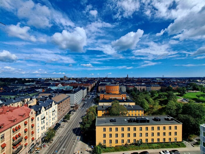 Stadsutsikt med blå himmel över bostadshus, väg och grönområden, med ett centralt placerat runt orangefärgat hus i mitten.