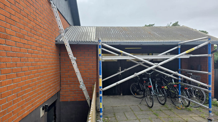 Stege lutande mot tegelvägg bredvid byggställning och plåttak över cykelparkering.
