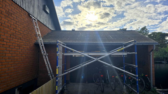 Byggarbetsplats med tak, ställning och stege mot röd tegelvägg, under en molnig himmel. Cyklar syns under ställningen.