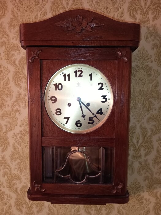 Mekaniskt väggur i mörkt trä med blomsterdekor, urtavla med svarta siffror och visare mot en blommig tapetbakgrund.