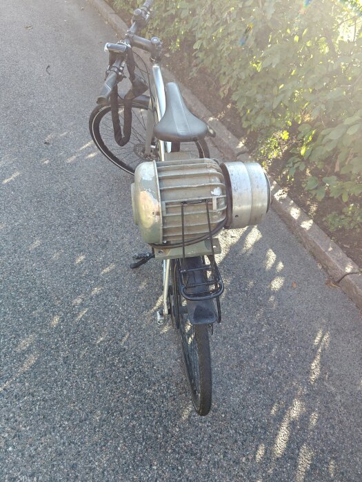 Cykel på trottoar med vintage motor fastsatt på pakethållaren, omgiven av grönska i morgonsol.
