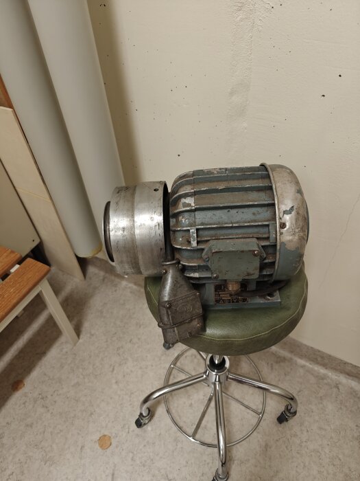 Gammal elektrisk motor placerad på en snurrstol i ett rum med vitmålad vägg.
