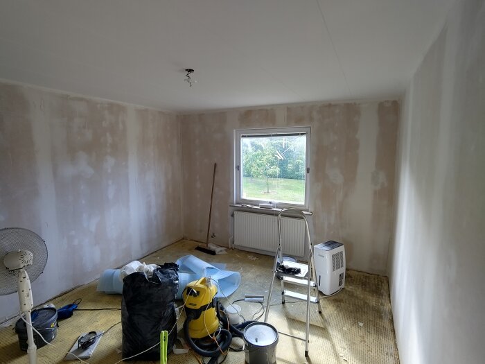 Rum under renovering, väggar täckta med glasfiberväv, stege vid fönster, dammsugare och verktyg spridda på golvet.