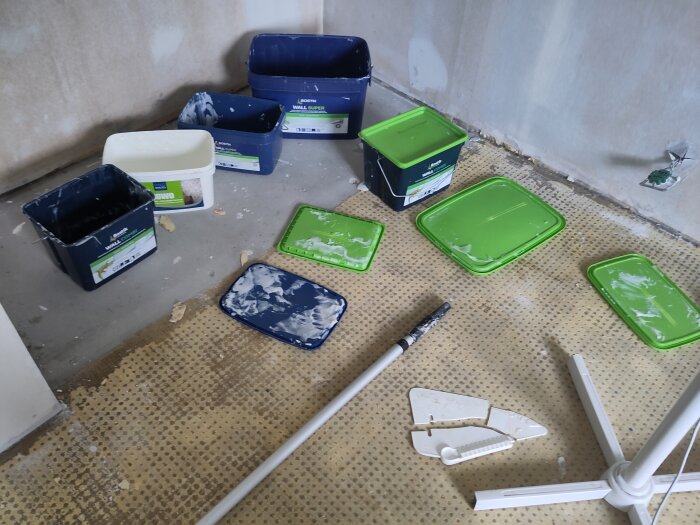 Plastbyttor med färgrester på ett golv, färgverktyg och en trasig tapetspade i ett renoveringsrum.