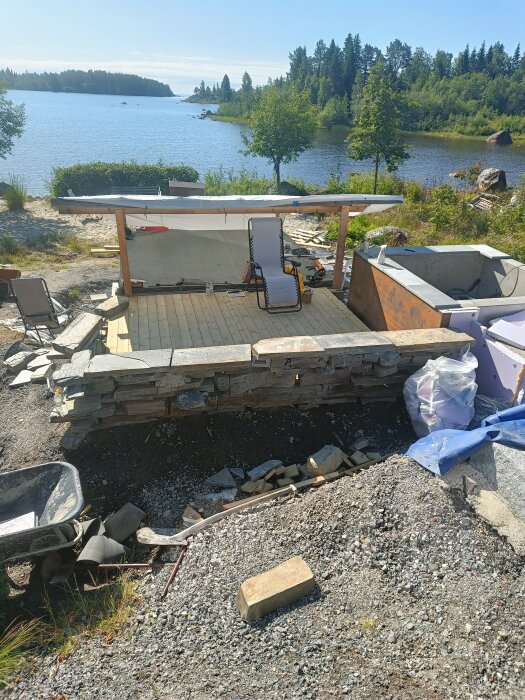 Stenmur, pooldäck och en halvfärdig pool vid vattnet, samt träd och skog i bakgrunden.