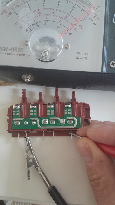 En hands testar kretsar på en demonterad röd och grön knappsats med en multimeter i bakgrunden.