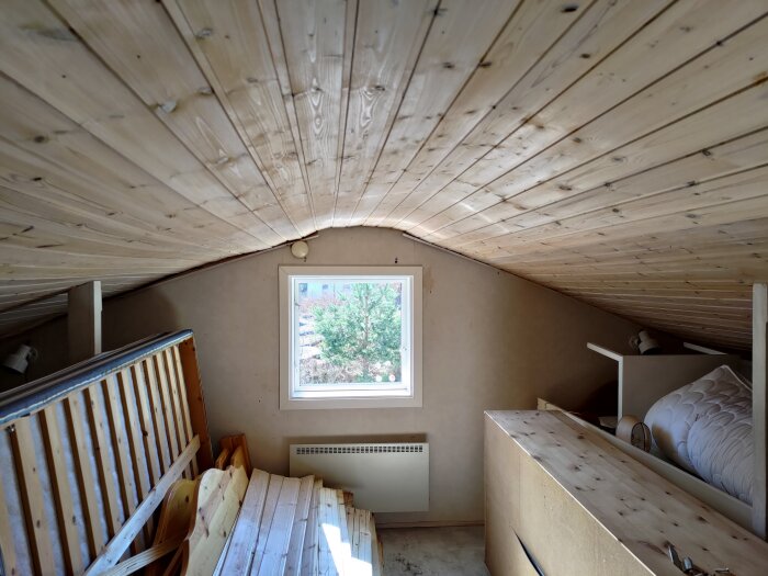 Trångt utrymme med snedtak, ett litet fönster och diverse trä- och byggmaterial staplat.