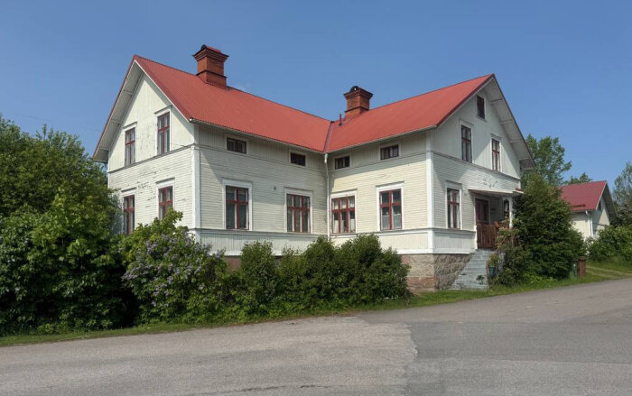 Stort vitmålat trähus i två våningar med rött plåttak, två skorstenar och omgiven av grönska, beläget intill en väg.